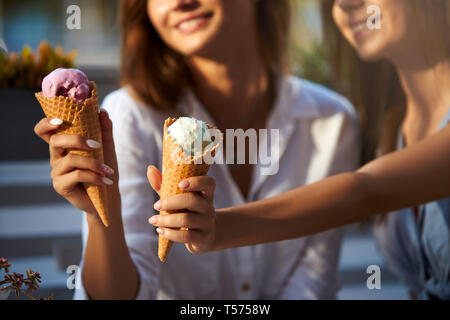Close up shot of ice cream cones in hand of a woman standing with her friend. Two young women outdoors eating icecream on a sunny day. Isolated view, 