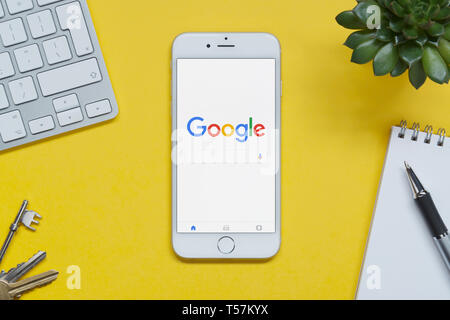 An iPhone showing the Google website rests on a yellow background table with a keyboard, keys, notepad and plant (Editorial use only). Stock Photo