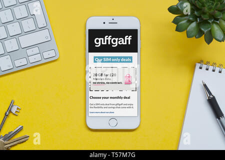 An iPhone showing the Giffgaff website rests on a yellow background table with a keyboard, keys, notepad and plant (Editorial use only). Stock Photo