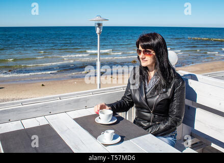 young brunette woman in black leather jacket and sunglasses drinking coffee in an  outdoor cafe near the sea on sunny day Stock Photo