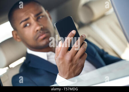 Young black businessman sitting in the car holding smartphone close-up using application blurred background looking aside concerned Stock Photo