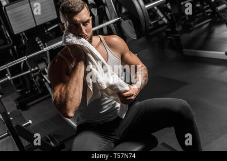 Bearded man bodybuilder sitting on bench at gym wiping off sweat from face with towel thoughtful Stock Photo