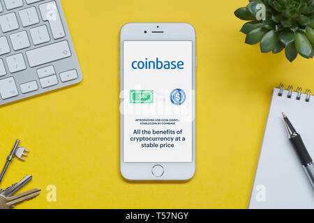An iPhone showing the Coinbase website rests on a yellow background table with a keyboard, keys, notepad and plant (Editorial use only). Stock Photo