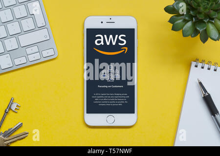 An iPhone showing the Amazon Web Services website rests on a yellow background table with a keyboard, keys, notepad and plant (Editorial use only). Stock Photo