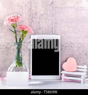 Home decor with blooming carnation and tablet as photo frame beside wall on the table - Close up, copy space, mock up, beautiful mothers day concept. Stock Photo
