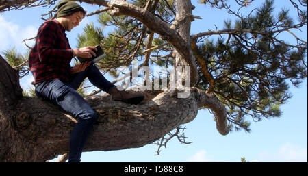 Shot of an authentic caucasian man sat on a tree branch reading a book and smiling. Stock Photo