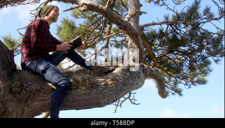 Shot of an authentic caucasian man sat on a tree branch day dreaming, reading a book and smiling. Stock Photo