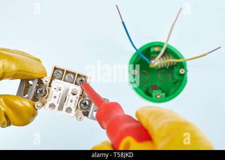 Electrician in yellow protective gloves is twisting screw in part of light switch by hand tool, close-up. Stock Photo