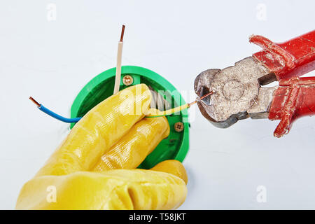 Electrician in yellow protective gloves is cutting ends of copper wiring of round electrical box for wall light switch with nippers tool. Stock Photo