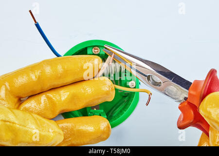 Worker in yellow protective gloves is cutting ends of copper wiring of round electrical box for wall light switch with long nose pliers. Stock Photo