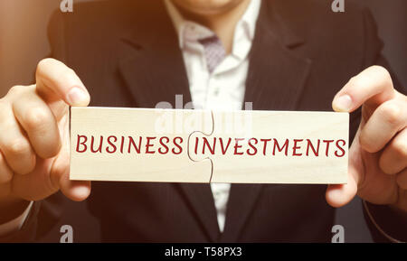 Businessman collects wooden puzzles with the words business investments. Increase capital. Investing your assets in your own or someone else's busines Stock Photo