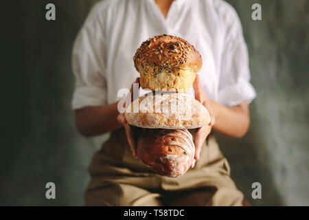 Close up of female baker holding different types of bread. Chef holding various types of loaf bread against gray background. Stock Photo