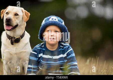 Smiling young boy sitting in a field next to his pet dog. Stock Photo