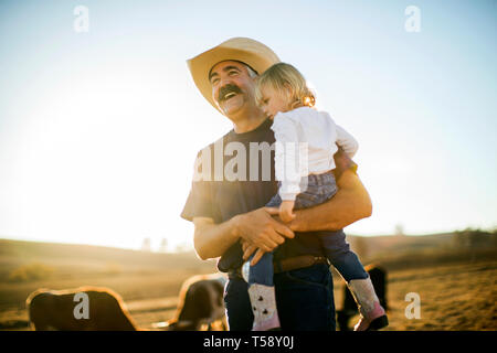 Cowboy rancher holding his toddler daughter in his arms while they are out on the ranch. Stock Photo