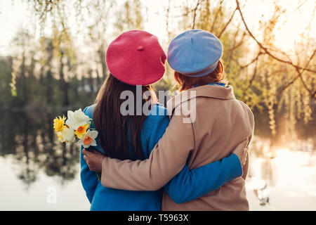 Happy senior mother with flowers and her adult daughter hugging by spring river at sunset. Mother's day concept. Family values Stock Photo