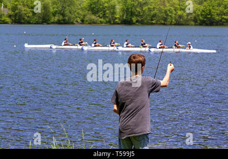 New Preston, CT USA. May 2012. A young man fishing at a lake despite the noise and cheering of a crew rowing race going on in front of him. Stock Photo
