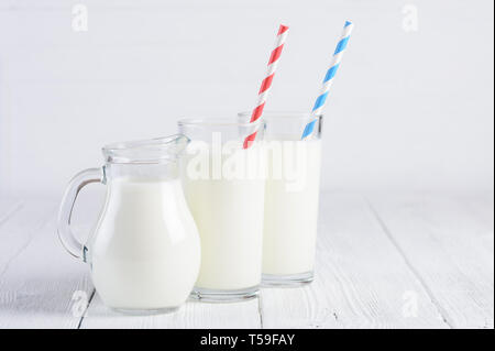 Glasses of milk with stripped blue and red paper straws and jug of milk on white wooden table still life Stock Photo