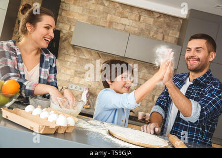 Mother father and son at home standing at table in kitchen together mom making dough watching dad giving high five to boy with floured hands smiling c Stock Photo