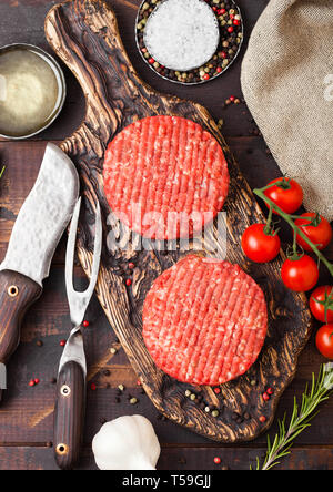 Fresh raw minced homemade farmers grill beef burgers on vintage chopping board with spices and herbs and fork with knife on wooden board. Stock Photo