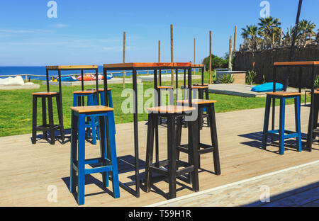 High table and chairs on wooden floor in exterior cafe restaurant with the beach and puffs, blue sky, clouds, trees and beautiful scenery view Stock Photo