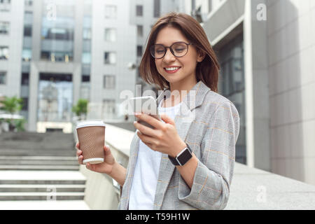 Productive morning. Young lady in eyeglasses standing on the city street with coffee reading message on smartphone cheerful Stock Photo