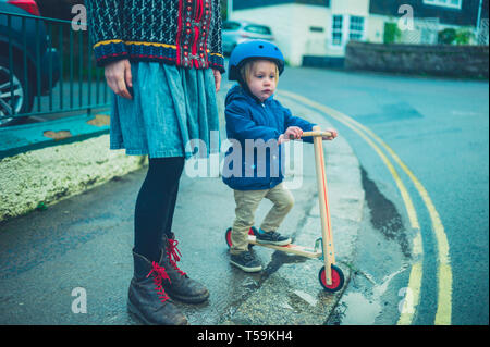 A mother is standing in the street with her toddler who is riding a scooter Stock Photo