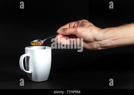 Male hand pouring granulated instant coffee on a white mug, isolated over black. Stock Photo