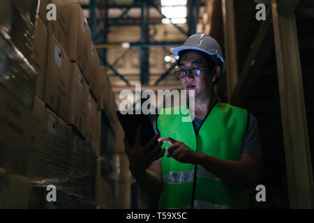 Young Asian male warehouse worker using a digital tablet inside warehouse to check inventory and stock management in factory Stock Photo
