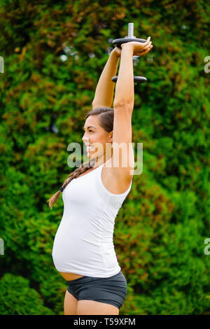 Pregnant Woman Exercising Triceps Extension With Dumbbell In Park Stock Photo