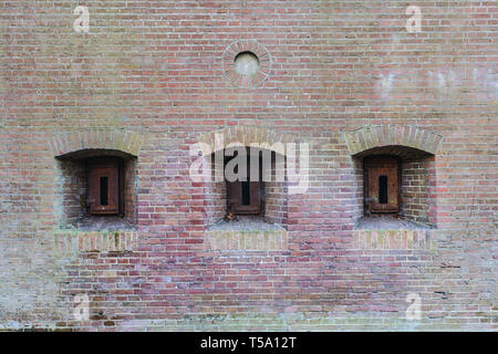 Brick wall of an old fortress with three embresures closed with rusty iron hatches. Stock Photo