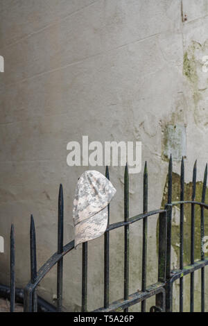 Abandoned grey and tan pixelated camouflage hat on a wrought iron fence gate to an alley with damaged stucco wall Stock Photo
