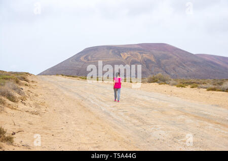 Woman in pink walking on a sandy path towards a volcano in La Graciosa island Stock Photo