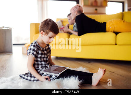 A small girl with mother and brother using tablet indoors on the floor at home. Stock Photo