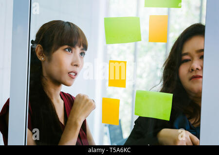 Close-up of serious and determined Asian business women looking at transparent glass board with sticky notes Stock Photo
