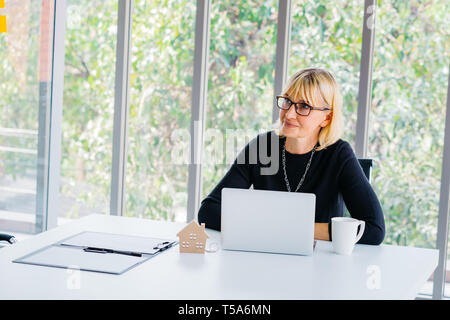 Serious Caucasian senior executive business woman in casual with laptop sitting in board room and looking away thinking - with copy space Stock Photo
