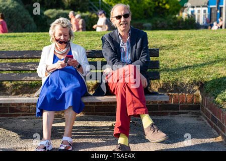 An elderly couple sitting together on a bench in the summer sun, she is using a mobile phone in Littlehampton in England Stock Photo
