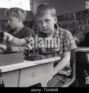 1960s, historical, a schoolboy sitting at his single wooden topped metal desk playing with a set of  wooden dominoes, England. Dominoes is a tile-based gams played with small rectangular 'domino' tiles, of which the end is marked with a number of spots or is blank. Dominoes are used in primary education as a a learning resource and are excellent for developing number sense and maths skills, as well as helping in counting, matching, sorting, classifying and other important skills. Stock Photo