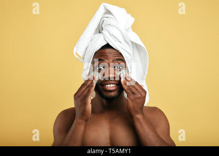 puzzled man with palms on his cheek and green cream posing to the camera, isolated yellow background. man is allergic to the cosmetic, skin problems Stock Photo
