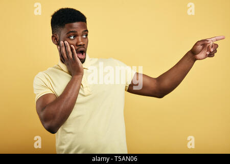 astonished emotional young dark-skinned man dressed in stylish T-shirt,touching his cheek, pointing at copy space on yellow wall for text. guy is afraid of somebody, close up portrait Stock Photo