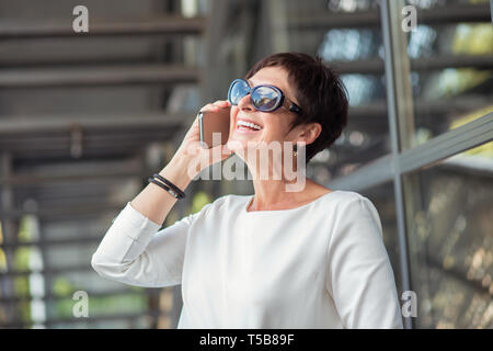 Trendy middle-aged mature woman wearing white blouse wearing black sunglasses reflecting clouds and skies is speaking, talking on smartphone cheerfull Stock Photo