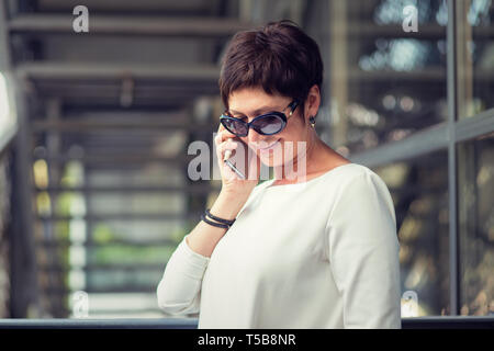 Talking on phone smiling. Side profile view portrait of happy mature lady using smart phone looking down urban metallic stairs on background. Model in Stock Photo