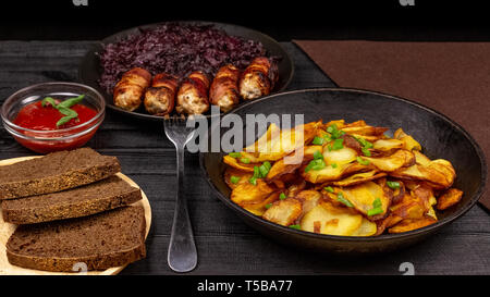 Roasted potato in the pot, pieces of rye black bread on a wooden serving plate, homemade sausages in bacon and stewed cabbage on a black rustic backgr Stock Photo