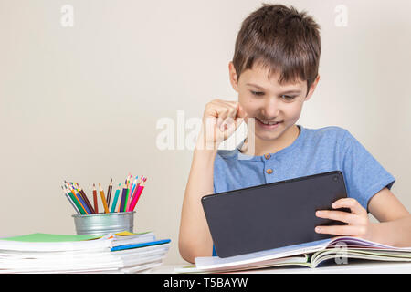 Kid sitting at table with books notebooks and playing games on tablet computer Stock Photo