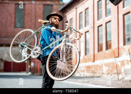 Lifestyle portrait of a bearded hipster dressed stylishly with hat and jacket carrying his retro bicycle on the urban background Stock Photo
