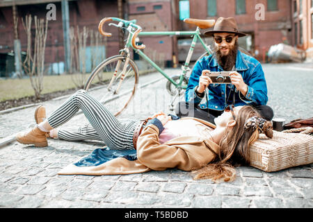 Stylish man photographing young woman lying on the street, having fun together on the urban background Stock Photo