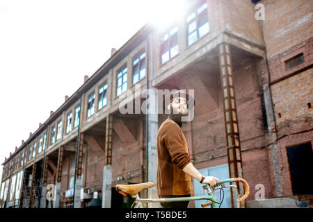 Lifestyle portrait of a bearded hipster dressed stylishly walking with retro bicycle on the industrial urban background