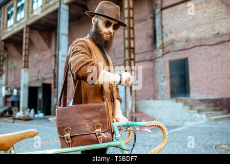 Lifestyle portrait of a bearded hipster dressed stylishly walking with retro bicycle on the industrial urban background Stock Photo