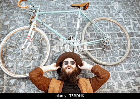 Portrait of a stylish bearded man in hat lying with retro bicycle on the pavement street, view from above Stock Photo