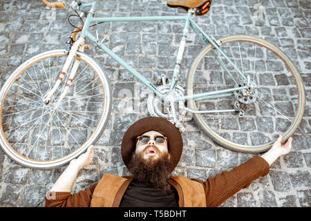 Portrait of a stylish bearded man in hat lying with retro bicycle on the pavement street, view from above Stock Photo