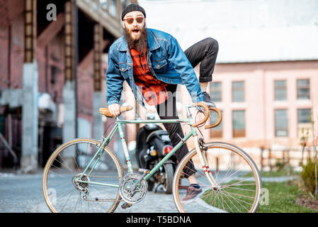 Portrait of a bearded man as a crazy hipster having fun with retro bicycle outdoors on the industrial urban background Stock Photo